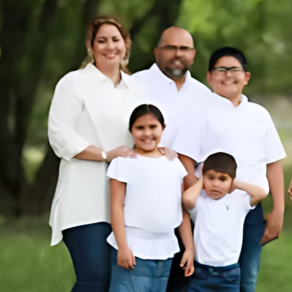 A smiling family of five, two adults and three children, posing outdoors in white shirts and blue jeans.