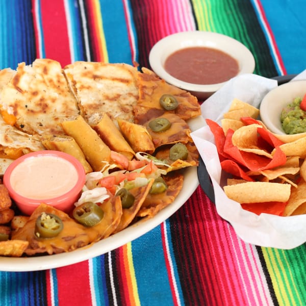 A platter of quesadillas, taquitos, nachos with jalapeños, and dipping sauces on a colorful striped blanket.