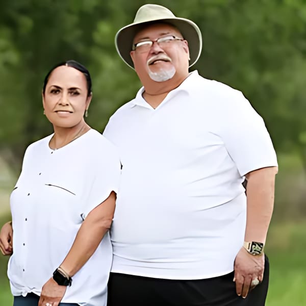 A man in a hat and a woman in white shirts stand together against a green background.