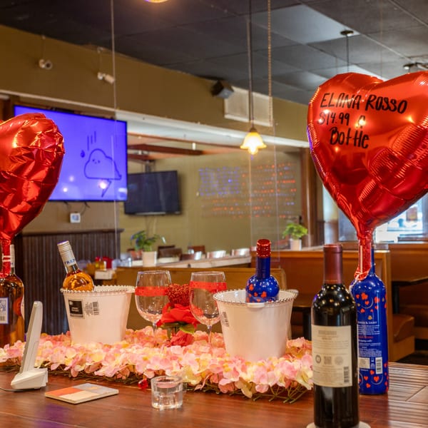 Table decorated with pink flowers, wine bottles, glasses, and heart-shaped balloons in a restaurant.
