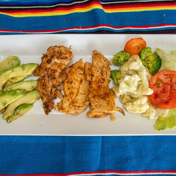 Grilled chicken breast with avocado slices, steamed broccoli, cauliflower, and sliced tomato on a white plate.